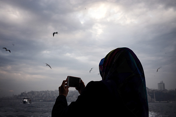 一名头戴头巾的年轻的穆斯林女性站在船舷外用手机拍摄海鸥.