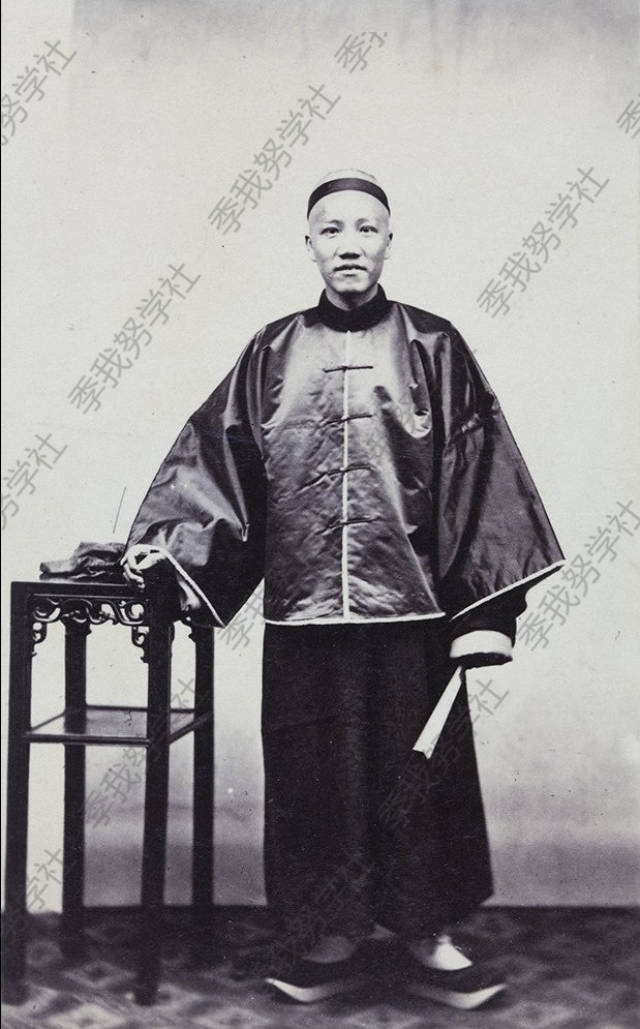 1870-1875年,一个身穿中式长袍马褂的中国男性.他手中拿着折扇站着.