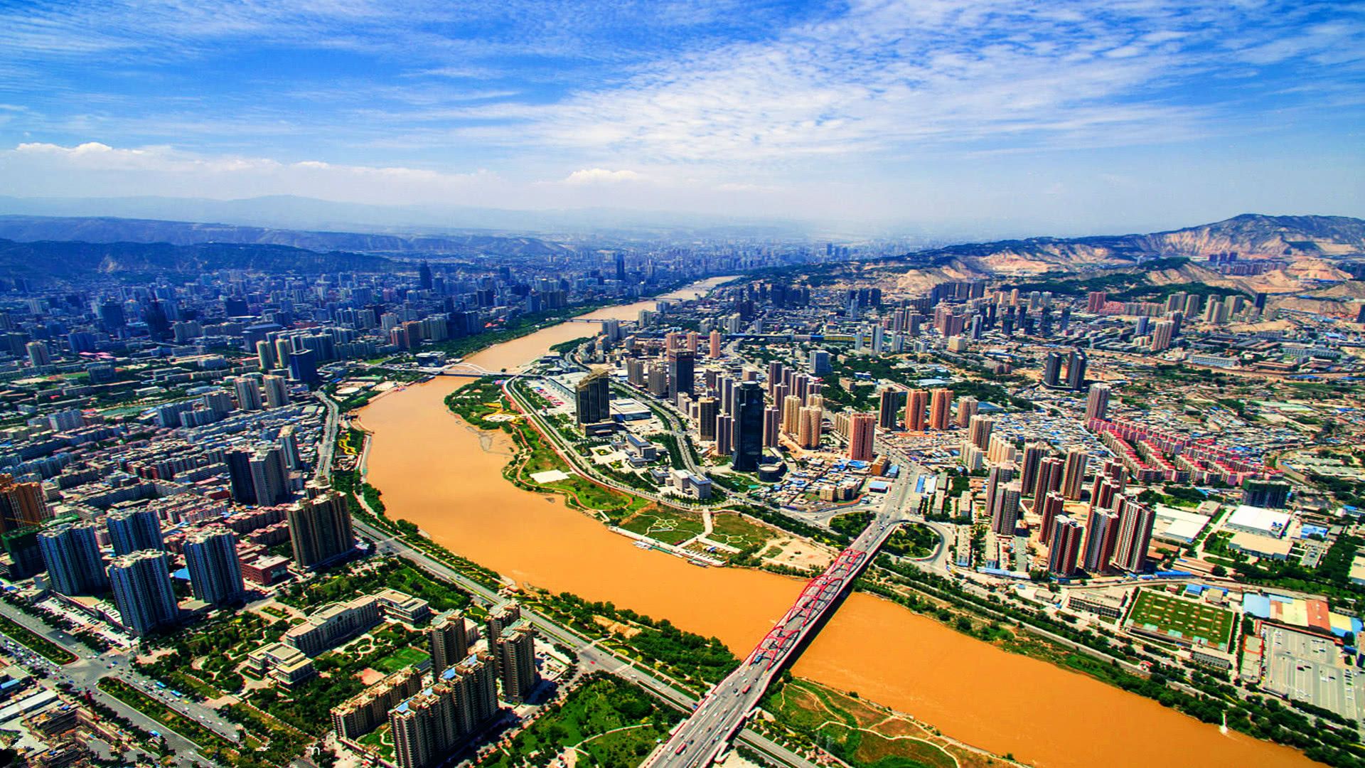 这是一张城市全景航拍照片，显示了蜿蜒的河流穿过城市中心，两岸高楼林立，大桥横跨河面，远处有山脉。