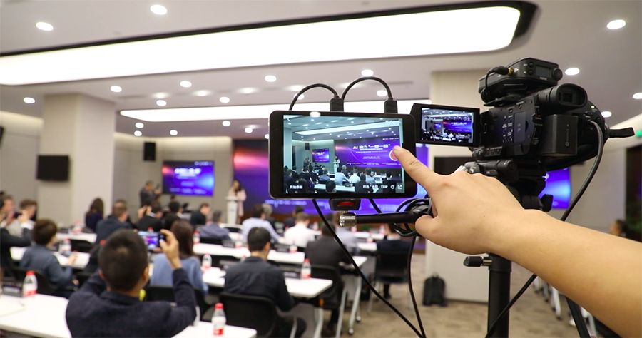 图片展示了一个会议室内的场景，前方有发言台，观众在听讲。一台摄像机正在录制发言人和听众。
