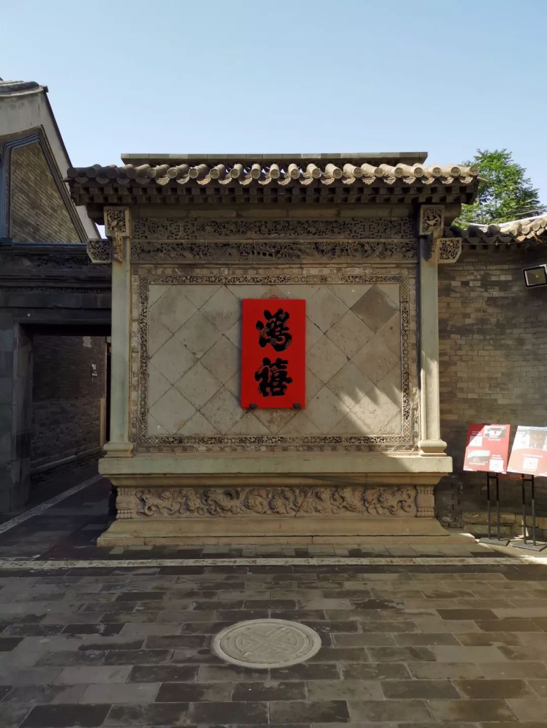 影壁,壁身为正方形,周围是砖雕装饰,中间挂有红底黑字的"鸿禧"木牌